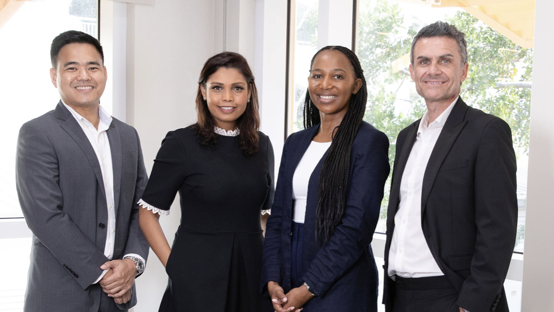 Four business people of different races stand in front of a glass window smiling at the camera wearing business wear